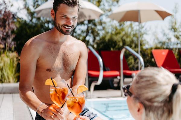 Ein entspanntes Paar genießt erfrischende Aperol Spritz Cocktails am Poolrand unter Sonnenschirmen. Die fröhliche Stimmung, warmes Sonnenlicht und leuchtende Farben schaffen ein sommerliches Urlaubsgefühl in angenehmer Atmosphäre.