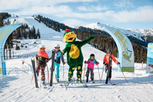 Vier Kinder posieren mit Skiausrüstung und einem fröhlichen Maskottchen im Drachenkostüm vor malerischer Winterlandschaft. Freudige Stimmung, klare Bergluft und beschneite Hänge sorgen für ein unvergessliches Skierlebnis im Familien-Skigebiet. Bunte Kleidung und lachende Gesichter laden zum Mitmachen ein.