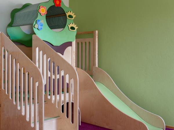 Ein moderner Indoor-Spielbereich mit einer kindgerechten Holzrutsche und einem bunten Kletterelement in Form eines Baumes. Die sanften Gr&uuml;n- und Holzt&ouml;ne schaffen eine freundliche Atmosph&auml;re. Ideal zum sicheren Spielen und Klettern f&uuml;r Kleinkinder in Innenr&auml;umen.