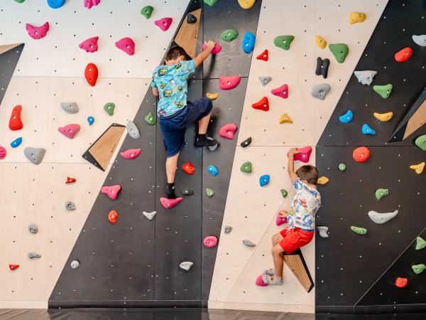Zwei Kinder klettern an einer bunten Boulderwand mit verschiedenen farbigen Griffen und Tritten. Die Wand bietet abwechslungsreiche Routen und eine spannende Herausforderung in einer sicheren Indoor-Umgebung. Perfekt zum Spielen, Ausprobieren und F&ouml;rdern motorischer F&auml;higkeiten.