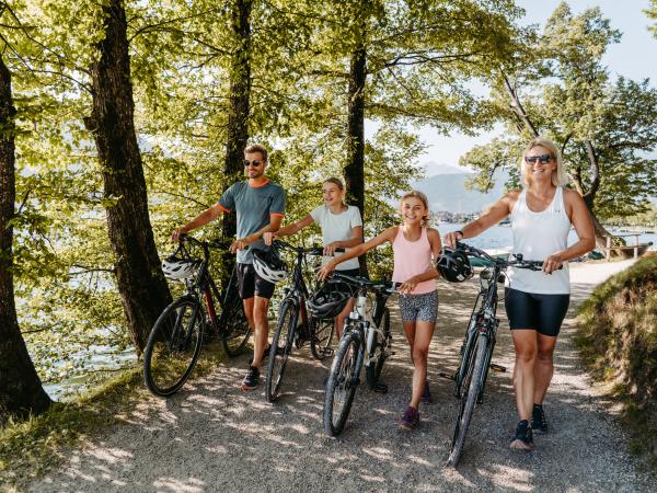 Eine fröhliche Familie genießt einen gemeinsamen Fahrradausflug entlang eines idyllischen Waldwegs am See. Umgeben von grünen Bäumen und sanftem Sonnenlicht erleben Groß und Klein naturnahe Bewegung und Spaß an der frischen Luft. Ideal für aktive Familien.