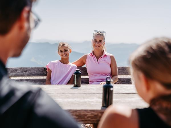 Eine lachende Mutter mit ihrem Kind genie&szlig;t den Tag auf einer Bergh&uuml;tte, w&auml;hrend Vater und Tochter ihnen gegen&uuml;ber sitzen. Im Hintergrund erstreckt sich eine beeindruckende Alpenlandschaft. Wasserflaschen auf dem Holztisch runden die gem&uuml;tliche Stimmung ab.