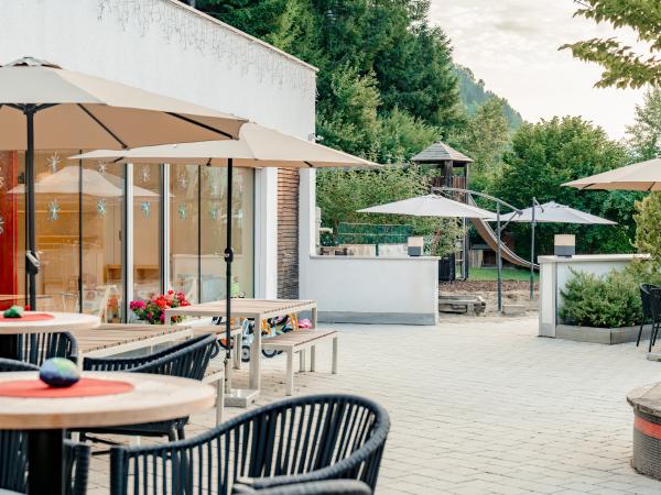 Die helle Sonnenterrasse bietet moderne Sitzgelegenheiten unter gro&szlig;en Sonnenschirmen und l&auml;dt zum entspannten Verweilen ein. Im Hintergrund sind ein Spielplatz und &uuml;ppiges Gr&uuml;n zu sehen &ndash; ideal f&uuml;r Familien und Genie&szlig;er. Die einladende Atmosph&auml;re sorgt f&uuml;r Erholung im Freien.
