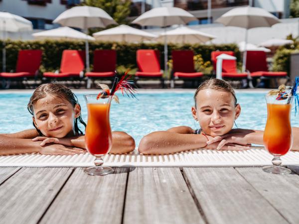 Zwei Kinder genie&szlig;en einen sonnigen Tag im Freibad und lehnen entspannt am Beckenrand. Vor ihnen stehen bunte, erfrischende Cocktails mit Deko, im Hintergrund sorgen rote Liegen und wei&szlig;e Sonnenschirme f&uuml;r Urlaubsstimmung. Die Szene vermittelt Sommer, Erholung und Lebensfreude.