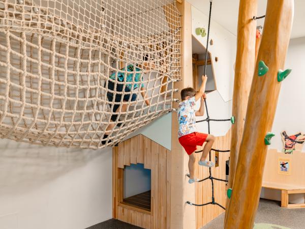 Zwei Kinder erkunden einen spannenden Indoor-Spielplatz mit Kletterelementen. Ein Junge erklimmt ein Kletternetz, während ein weiteres Kind eine Kletterwand und ein Seil nutzt. Helle Holzelemente und freundliche Farben sorgen für ein einladendes Ambiente – ideal für junge Abenteurer.