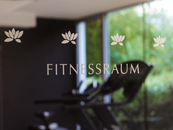 Ein heller Fitnessraum mit gro&szlig;en Fenstern, die den Blick ins Gr&uuml;ne erm&ouml;glichen. Im Vordergrund befindet sich Fitness-Equipment, das zur aktiven Bewegung einl&auml;dt. Die Glaswand ist mit Lotusbl&uuml;ten dekoriert und schafft eine motivierende, entspannte Atmosph&auml;re f&uuml;r das Training.