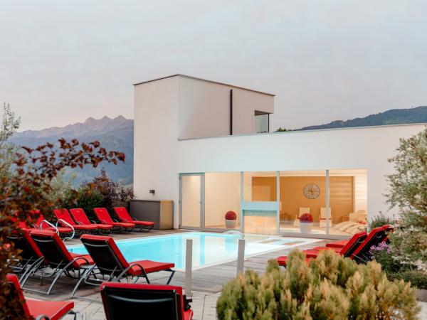 Ein stilvoll gestalteter Au&szlig;enpool mit eleganten roten Liegest&uuml;hlen l&auml;dt zum Entspannen ein. Im Hintergrund beeindruckt die alpine Berglandschaft, w&auml;hrend das moderne Geb&auml;ude mit gro&szlig;en Glasfronten f&uuml;r eine helle und einladende Atmosph&auml;re sorgt. Ideal f&uuml;r erholsame Stunden im Freien.