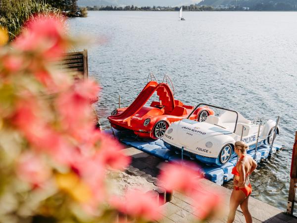 Zwei auff&auml;llige Tretboote im Sportwagendesign, eines rot mit Rutsche und eines wei&szlig;, liegen an einem Steg am ruhigen Seeufer. Im Vordergrund bunte Blumen, am Steg eine Person in Badebekleidung. Die Szene vermittelt Sommerstimmung und Freizeitspa&szlig; am Wasser.