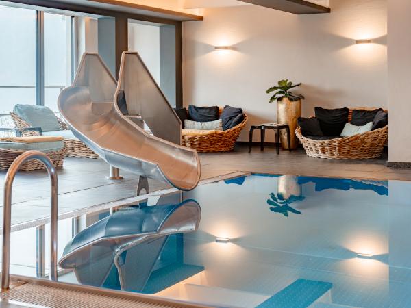 Ein stilvoller Innenpool mit einer glänzenden Edelstahlrutsche sorgt für Spaß und Entspannung. Im Hintergrund laden gemütliche Loungesessel und sanfte Beleuchtung zum Verweilen ein. Die klare Wasseroberfläche spiegelt die freundliche Atmosphäre des Wellnessbereichs wider.