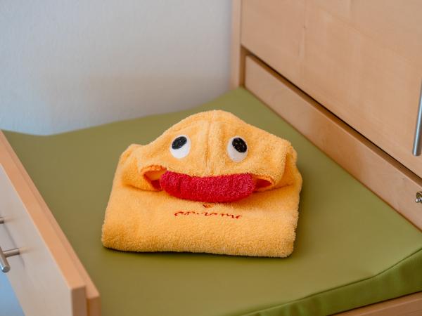 Ein gelbes Kinderhandtuch, liebevoll als Ente mit großen, aufgenähten Augen und rotem Schnabel gefaltet, liegt auf einer grünen Wickelauflage. Das Handtuch sorgt für fröhliche Stimmung und eignet sich ideal für Familien mit kleinen Kindern.