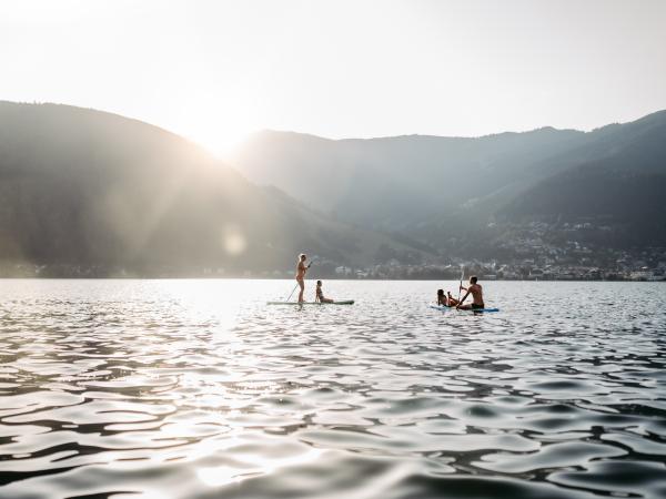 Zwei Personen genie&szlig;en Stand-Up-Paddling auf einem ruhigen See vor der Kulisse sanfter Berge bei Sonnenuntergang. Die Sonnenstrahlen spiegeln sich im Wasser, w&auml;hrend im Hintergrund sanfte H&uuml;gel und ein kleines Dorf zu sehen sind. Eine entspannte, naturnahe Atmosph&auml;re.