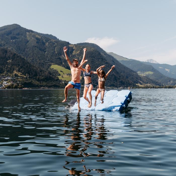 Eine fr&ouml;hliche Familie springt gemeinsam von einer Schwimminsel ins klare Wasser eines Alpensees. Im Hintergrund ragen bewaldete Berge auf und ein Segelboot liegt ruhig vor Anker. Die Szene strahlt Lebensfreude und Sommerurlaubsstimmung in idyllischer Naturkulisse aus.