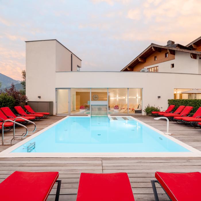 Moderner Außenpool mit klaren Linien, umgeben von stilvollen roten Liegestühlen auf Holzterrasse. Die Poolanlage lädt zum Entspannen ein und bietet einen beeindruckenden Blick auf die Alpenlandschaft. Ideal für individuelle Auszeiten und ruhige Wohlfühlmomente im familiengeführten Hotel.