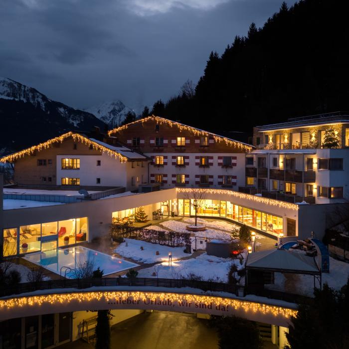 Gem&uuml;tliches Hotel im Alpenstil, festlich beleuchtet in der verschneiten Abendd&auml;mmerung. Die warmen Lichter schaffen eine einladende Atmosph&auml;re, w&auml;hrend im Hintergrund majest&auml;tische Berge das Bild abrunden. Perfekt f&uuml;r entspannte Winterurlaube in malerischer Kulisse.
