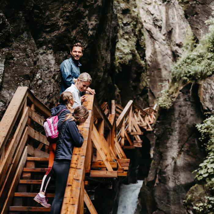 Eine Familie erkundet die beeindruckende Sigmund Thun Klamm auf hölzernen Stegen, die sich entlang steiler Felsen und über sprudelnde Wasserfälle schlängeln. Das Bild vermittelt Abenteuerlust und Naturnähe in einer alpinen Berglandschaft, ideal für Wanderbegeisterte und Naturliebhaber.