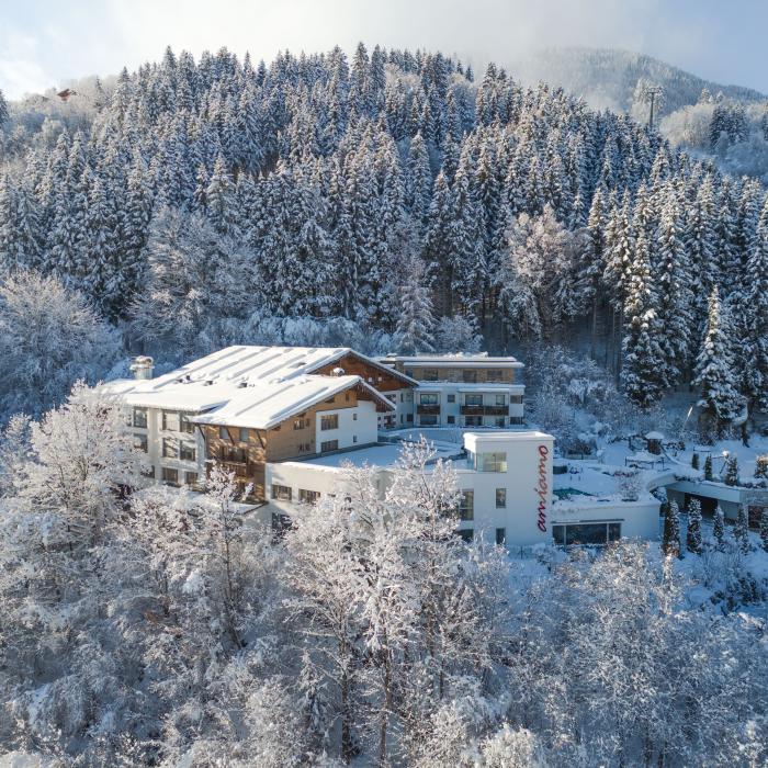 Das Bild zeigt ein einladendes Hotel, umgeben von schneebedeckten B&auml;umen am Waldrand. Die winterliche Atmosph&auml;re schafft eine idyllische Kulisse, w&auml;hrend das Geb&auml;ude harmonisch in die Landschaft eingebettet ist. Perfekt f&uuml;r einen erholsamen Winterurlaub in den Alpen.