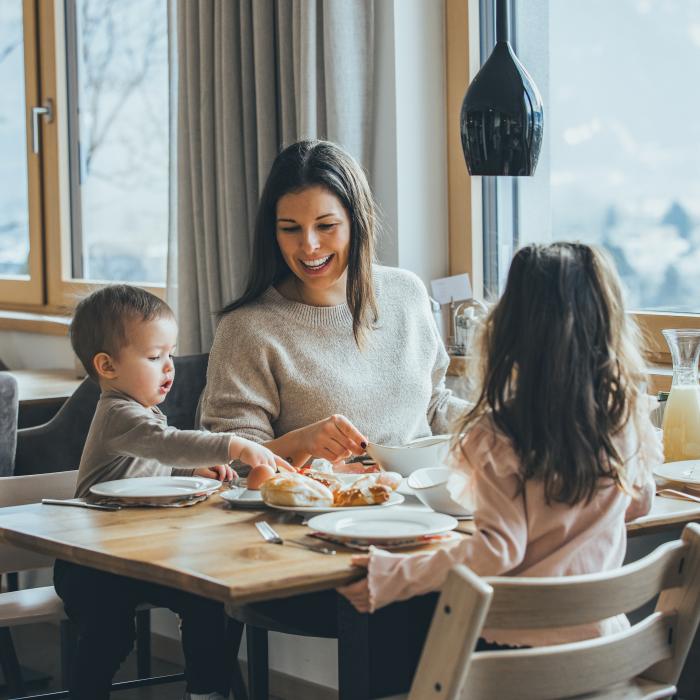 Eine fr&ouml;hliche Mutter genie&szlig;t gemeinsam mit ihren zwei Kindern ein gem&uuml;tliches Fr&uuml;hst&uuml;ck an einem Holztisch neben gro&szlig;en Fenstern. Nat&uuml;rliches Licht und eine sch&ouml;ne Aussicht auf die winterliche Landschaft schaffen eine harmonische, einladende Atmosph&auml;re.