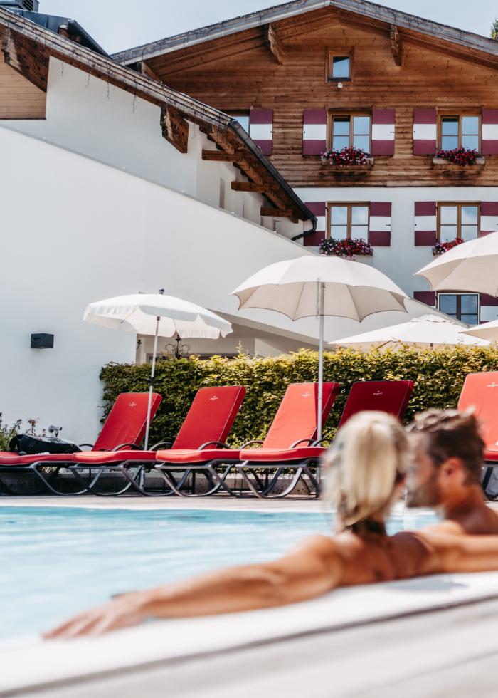 Ein Paar genießt einen sonnigen Tag im beheizten Außenpool eines gehobenen Resorts. Im Hintergrund sorgen rote Liegestühle und weiße Sonnenschirme vor einer charmanten Alpenkulisse für Urlaubsatmosphäre und ein Gefühl von Erholung und Wohlbefinden.