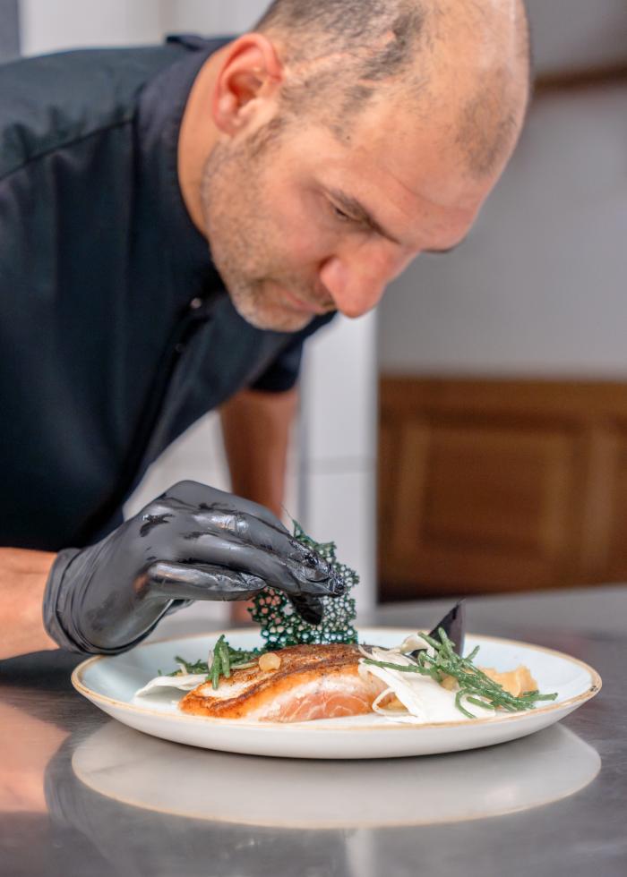 Ein Koch mit schwarzem Handschuh richtet ein kunstvoll dekoriertes Gourmetgericht an, konzentriert und mit Liebe zum Detail. Auf einem eleganten Teller werden frische Zutaten und feine Dekorationen stilvoll arrangiert, um ein kulinarisches Highlight zu schaffen. Perfekt für Genießer und Liebhaber gehobener Küche.