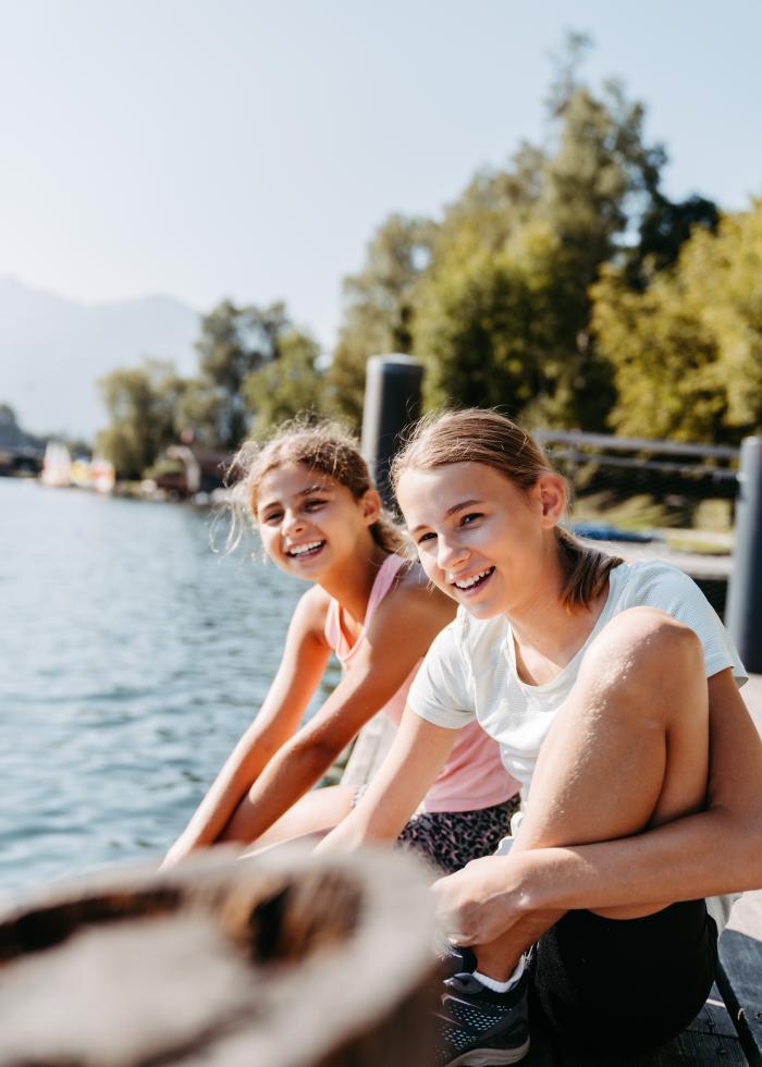 Zwei lachende M&auml;dchen sitzen entspannt an einem Holzsteg am klaren Seeufer. Umgeben von gr&uuml;nen B&auml;umen und sanften Bergen strahlen sie Freude und Sommerlaune aus. Das warme Sonnenlicht sorgt f&uuml;r eine lebendige, freundliche Stimmung in idyllischer Naturkulisse.