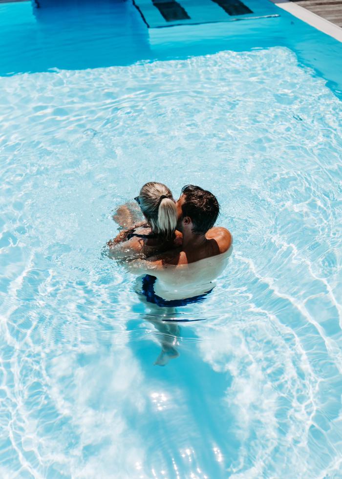 Ein verliebtes Paar umarmt sich liebevoll in einem klaren Au&szlig;enpool bei strahlendem Sonnenschein. Das Wasser schimmert t&uuml;rkisblau, w&auml;hrend Sonnenstrahlen f&uuml;r lebendige Reflexionen sorgen. Die entspannte Atmosph&auml;re l&auml;dt zum Verweilen und Wohlf&uuml;hlen ein &ndash; ideale Kulisse f&uuml;r eine Auszeit zu zweit.