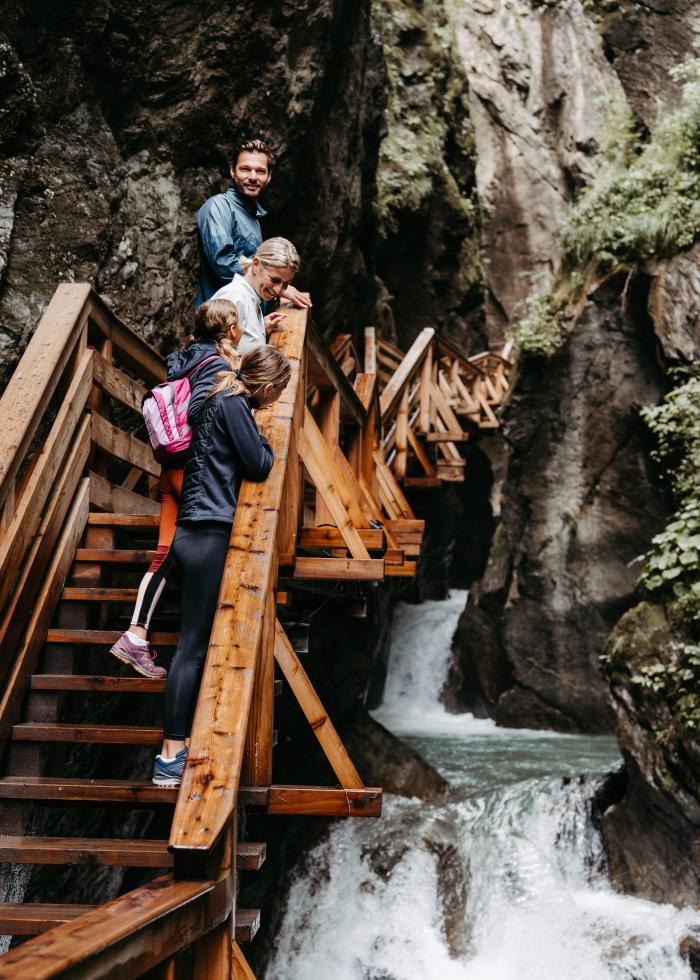 Eine Familie erkundet die beeindruckende Sigmund Thun Klamm auf hölzernen Stegen, die sich entlang steiler Felsen und über sprudelnde Wasserfälle schlängeln. Das Bild vermittelt Abenteuerlust und Naturnähe in einer alpinen Berglandschaft, ideal für Wanderbegeisterte und Naturliebhaber.