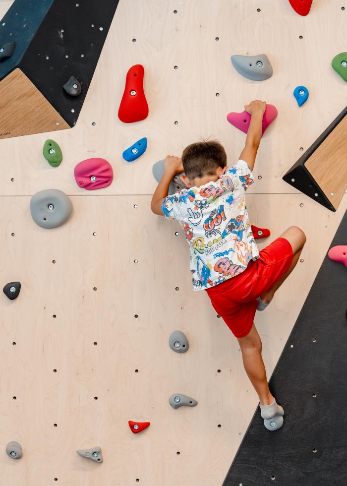 Ein Junge mit farbenfrohem T-Shirt und roten Shorts klettert an einer modernen Kletterwand mit bunten Griffen. Die Indoor-Kletterhalle bietet abwechslungsreiche Routen und sichere Bedingungen für junge Kletterbegeisterte. Die Szene vermittelt Bewegung, Konzentration und Freude am sportlichen Abenteuer.