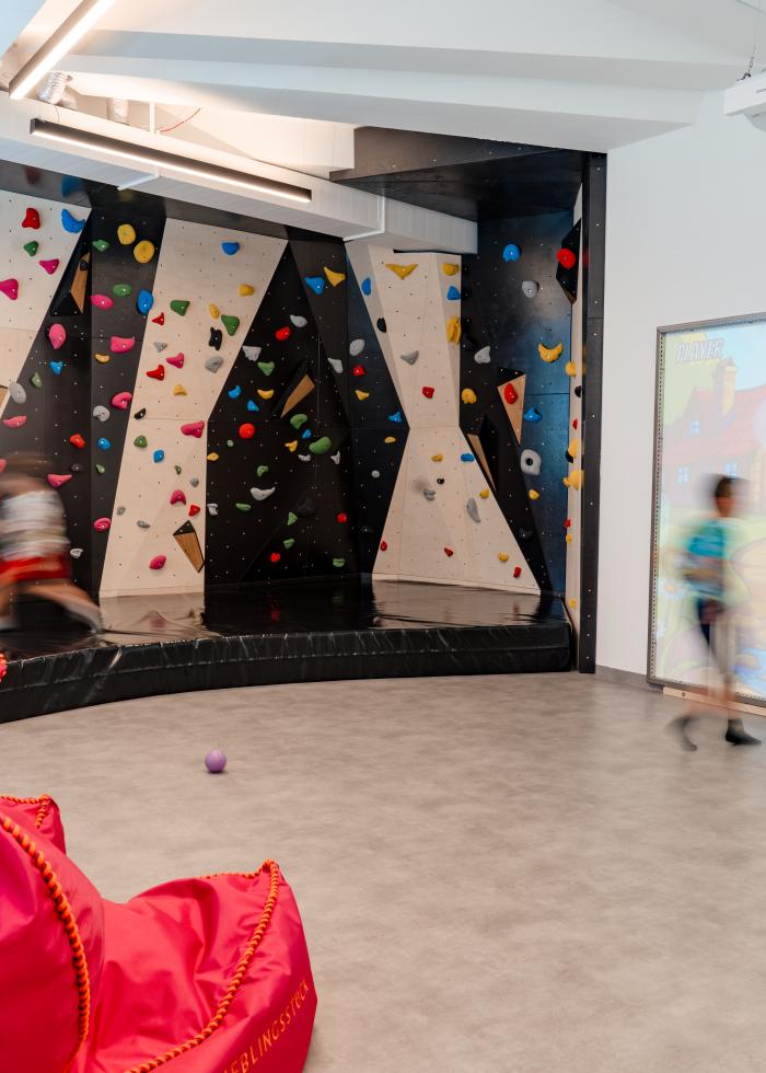 Ein heller Indoor-Spielraum mit einer farbenfrohen Kletterwand und gro&szlig;er interaktiver Projektionsfl&auml;che. Zwei Kinder sind im Raum aktiv, w&auml;hrend auff&auml;llige pinkfarbene Sitzs&auml;cke eine gem&uuml;tliche Ecke bilden. Die Kombination aus Bewegung und multimedialer Unterhaltung bietet vielseitigen Spielspa&szlig;.