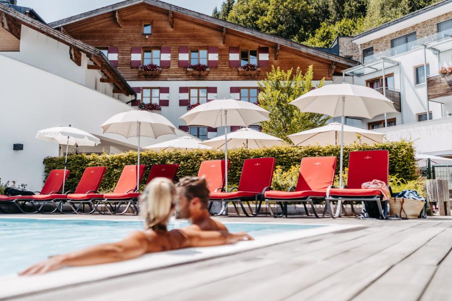 Ein Paar genießt einen sonnigen Tag im beheizten Außenpool eines gehobenen Resorts. Im Hintergrund sorgen rote Liegestühle und weiße Sonnenschirme vor einer charmanten Alpenkulisse für Urlaubsatmosphäre und ein Gefühl von Erholung und Wohlbefinden.