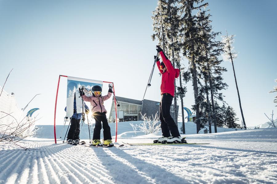 Ein Kind lernt mit Unterstützung eines Erwachsenen das Skifahren auf einer sonnigen, frisch präparierten Piste. Die Szene zeigt Spaß und gemeinsame Aktivität im familienfreundlichen Skigebiet, eingerahmt von verschneiten Bäumen und klarer Winterluft.