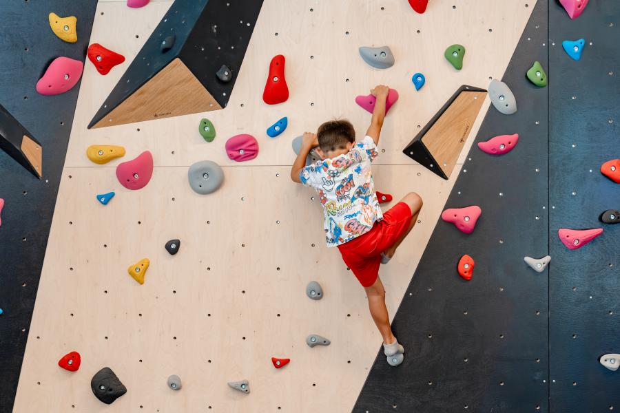 Ein Junge mit farbenfrohem T-Shirt und roten Shorts klettert an einer modernen Kletterwand mit bunten Griffen. Die Indoor-Kletterhalle bietet abwechslungsreiche Routen und sichere Bedingungen für junge Kletterbegeisterte. Die Szene vermittelt Bewegung, Konzentration und Freude am sportlichen Abenteuer.