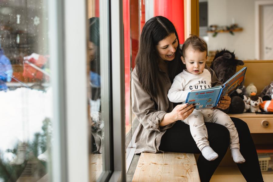 Eine liebevolle Szene zeigt eine junge Frau, die mit einem kleinen Kind auf einer Bank sitzt und gemeinsam ein Bilderbuch liest. Tageslicht f&auml;llt durch das gro&szlig;e Fenster und schafft eine warme, gem&uuml;tliche Atmosph&auml;re. Im Hintergrund ist ein freundlich eingerichteter Raum mit Spielzeug zu sehen.
