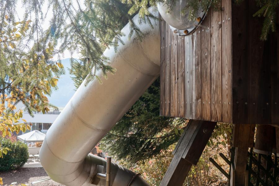 Eine große, silberne Rutsche führt aus einem hölzernen Baumhaus und schlängelt sich durch grüne Baumwipfel nach unten. Sonnenlicht glänzt durch das Blätterdach und verleiht der Szenerie eine verspielte und einladende Atmosphäre. Perfekt für ein Abenteuer im Freien.