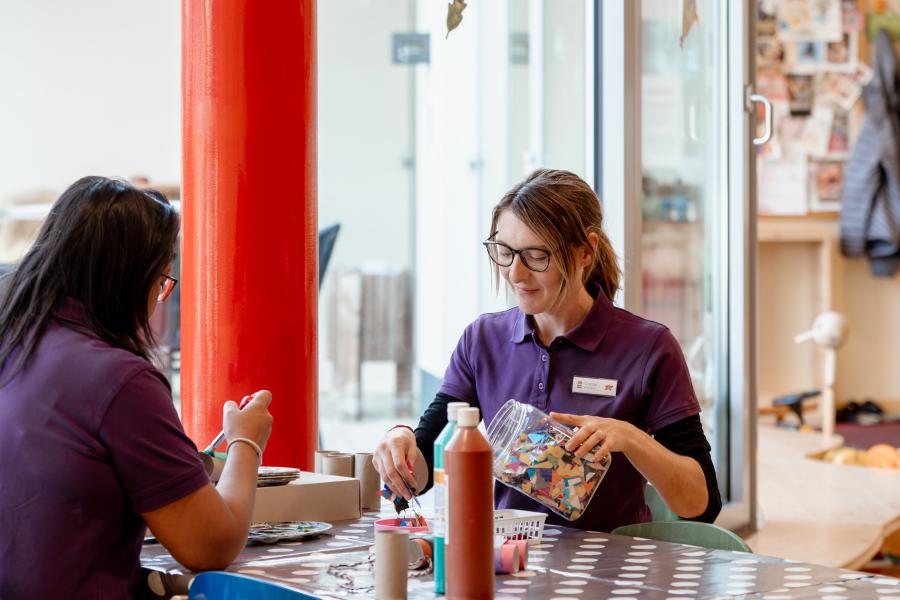 Zwei Betreuungspersonen sitzen an einem bunt gedeckten Tisch und basteln gemeinsam mit Farben und Materialien. Die gem&uuml;tliche Atmosph&auml;re wird durch herbstliche Fensterdekoration und warme Lichtverh&auml;ltnisse unterstrichen. Ein liebevoll gestalteter Raum f&ouml;rdert die Kreativit&auml;t der Kinder.