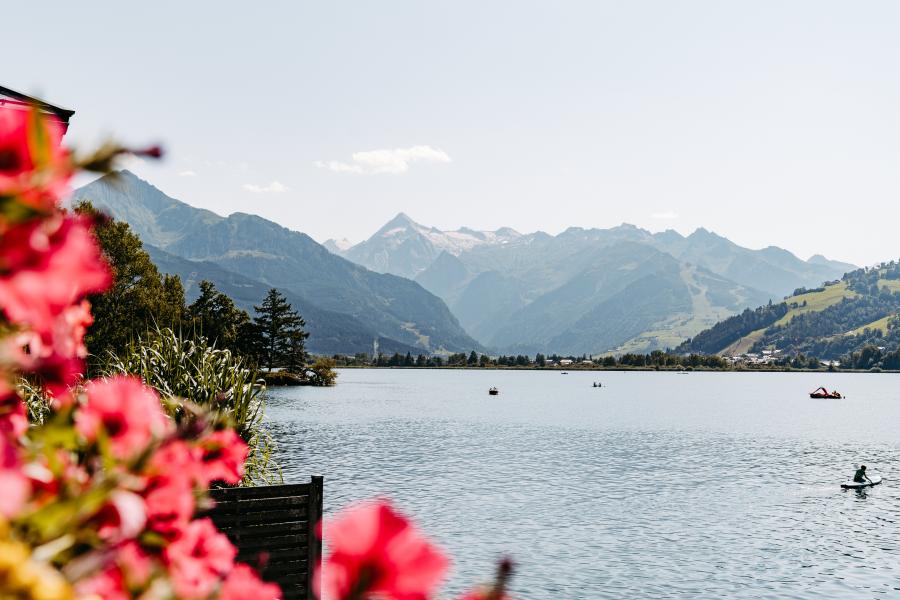 Ein idyllischer Blick &uuml;ber den Zeller See mit leuchtenden Blumen im Vordergrund und imposanten Alpen im Hintergrund. Das klare Wasser l&auml;dt zum Entspannen ein, w&auml;hrend Boote die ruhige Szenerie beleben. Perfekte Kulisse f&uuml;r Natur- und Outdoorliebhaber.