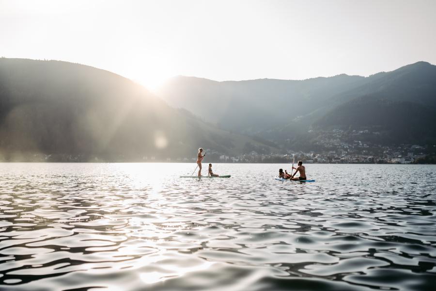 Zwei Personen genie&szlig;en Stand-Up-Paddling auf einem ruhigen See vor der Kulisse sanfter Berge bei Sonnenuntergang. Die Sonnenstrahlen spiegeln sich im Wasser, w&auml;hrend im Hintergrund sanfte H&uuml;gel und ein kleines Dorf zu sehen sind. Eine entspannte, naturnahe Atmosph&auml;re.