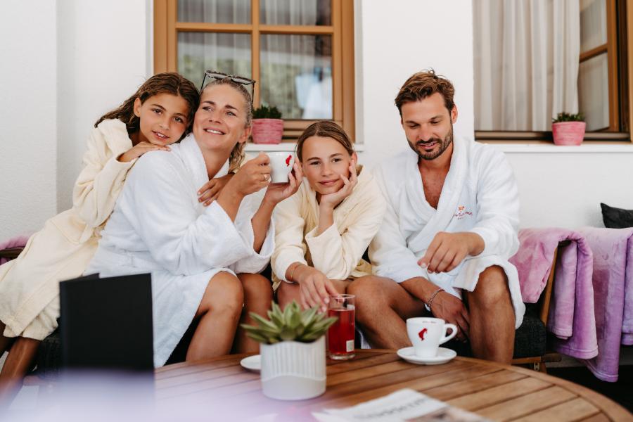 Eine glückliche Familie genießt gemeinsam Zeit auf einer sonnigen Terrasse. Alle tragen gemütliche Bademäntel, trinken heiße Getränke und schauen entspannt in die Kamera. Die gemütliche Atmosphäre wird durch Pflanzen und sanfte Farben im Hintergrund unterstrichen.
