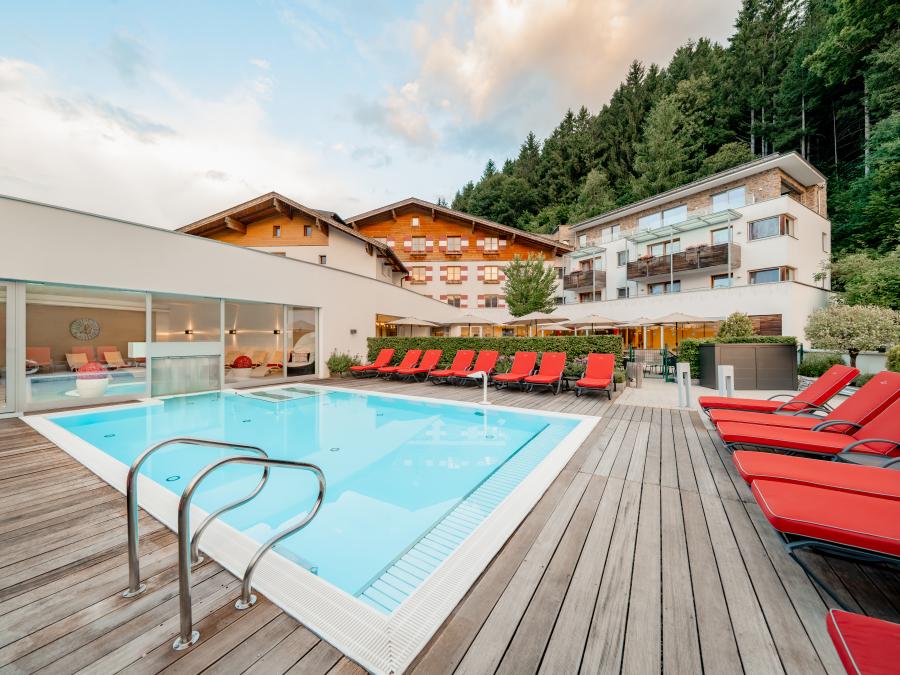 Moderner Au&szlig;enpool mit breiter Holzterrasse und stilvollen roten Liegest&uuml;hlen im Vordergrund. Im Hintergrund befindet sich ein elegantes Hotelgeb&auml;ude, umgeben von gr&uuml;nen B&auml;umen am Waldrand. Klare Linien und warme Farben schaffen eine entspannte Urlaubsatmosph&auml;re.